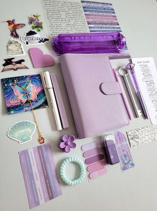Purple and lilac aesthetic bookish holiday gift bundle flatlay featuring a lavender planner cover, transparent purple pencil case, book tracker, pastel purple pens, stickers, bookmarks, scrunchie, tabs, and cute stationery accessories. A cozy Christmas gift set for book lovers, journalers, and students.