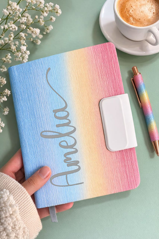 Pastel rainbow notebook with 'Amelia' on a light green surface with flowers and a cup of coffee.